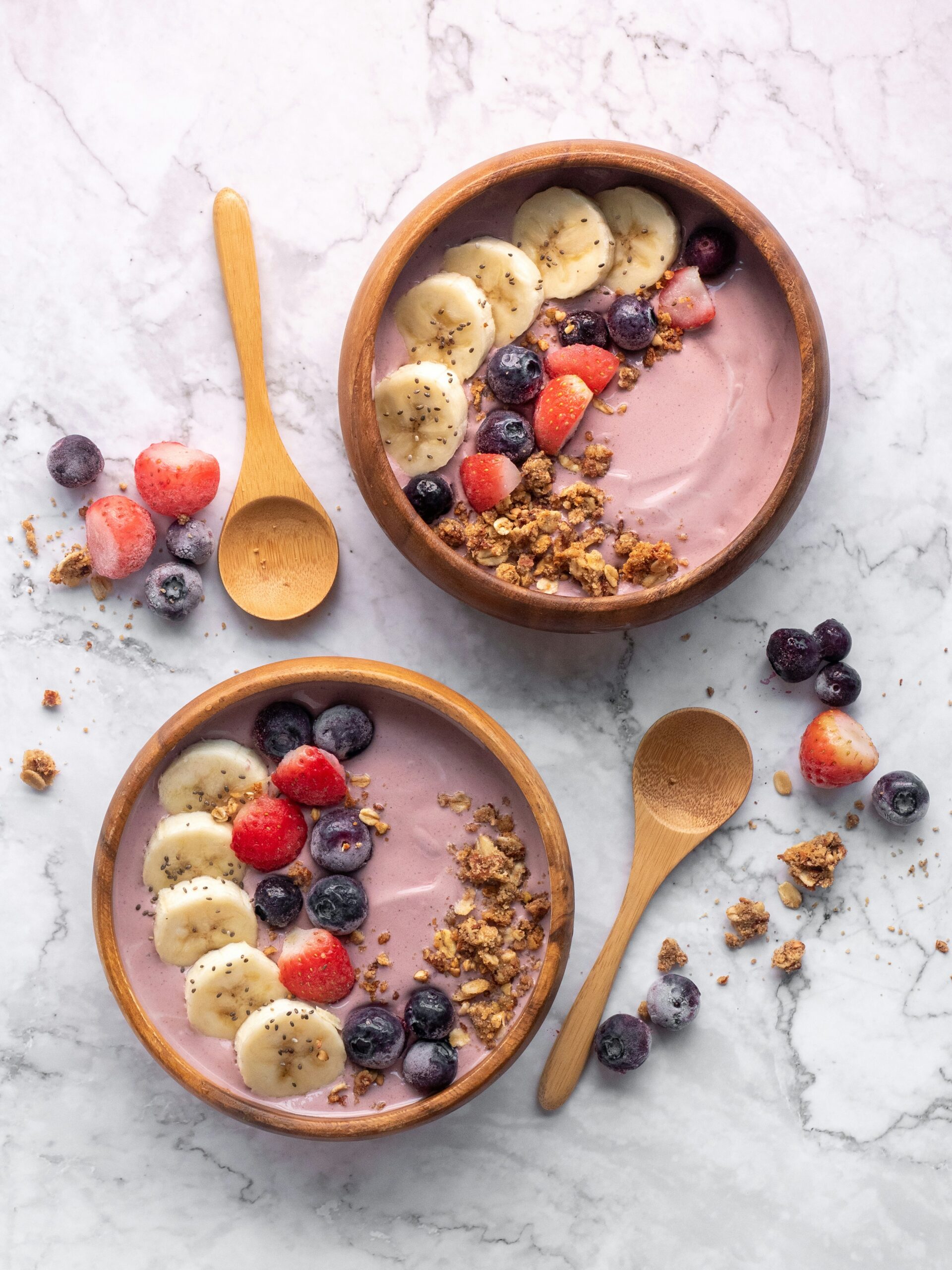2 vibrant colored smoothie bowls with banana, granola, and berries inside wooden bowls